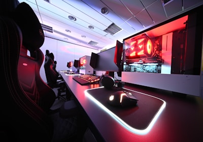 A row of high-end gaming computers on sleek desks, featuring RGB lighting and gaming accessories. The computers are housed in transparent cases with visible components, emitting a red glow. Comfortable gaming chairs with ergonomic design are positioned next to the desks, and the room's ceiling is modern with recessed lighting.