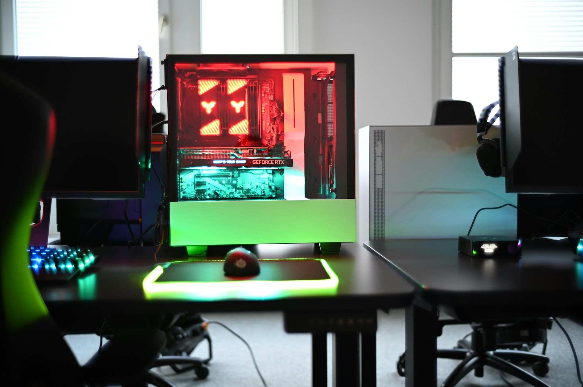 A modern gaming setup is displayed on a desk with multiple computer monitors and an open gaming PC tower. The tower is illuminated with red and green LED lights, showcasing a 'GeForce RTX' graphics card. A gaming mouse rests on a mousepad with glowing edges, and there is a keyboard with vibrant backlighting. The room appears well-lit with visible windows in the background.