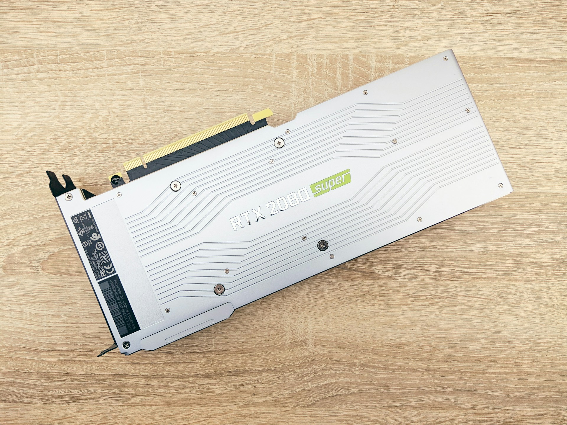 A graphics card is placed on a wooden surface with a minimalist design featuring a metallic silver finish. Engraved details include the text 'RTX 2080 SUPER' with an accented green 'super' logo. Visible are connector ports and screws, indicating parts of the card's functional design.