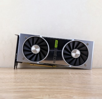 A metallic computer graphics card rests on a wooden surface. It features two large black cooling fans and a label indicating 'RTX 2080 Super'. The card has a sleek, modern design with silver and black elements.