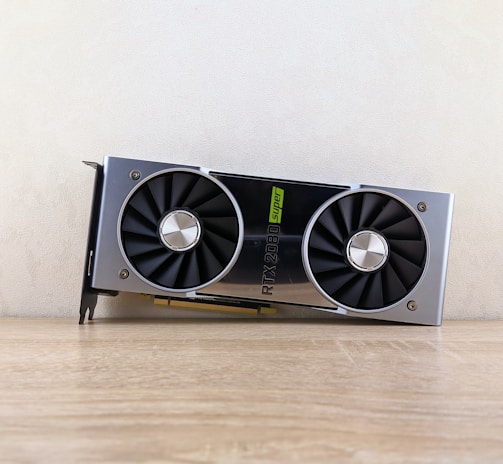 A metallic computer graphics card rests on a wooden surface. It features two large black cooling fans and a label indicating 'RTX 2080 Super'. The card has a sleek, modern design with silver and black elements.