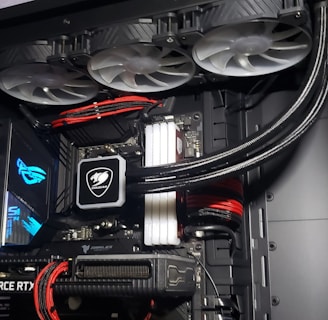 A high-performance gaming computer setup showing the interior components, including a GeForce RTX graphics card, cooling system with three fans, a motherboard, and visible wiring with red and black cables.