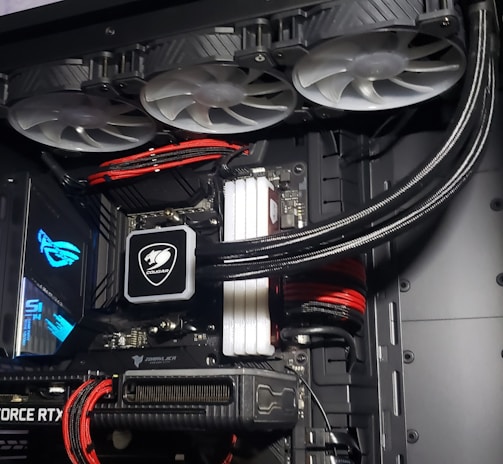 A high-performance gaming computer setup showing the interior components, including a GeForce RTX graphics card, cooling system with three fans, a motherboard, and visible wiring with red and black cables.