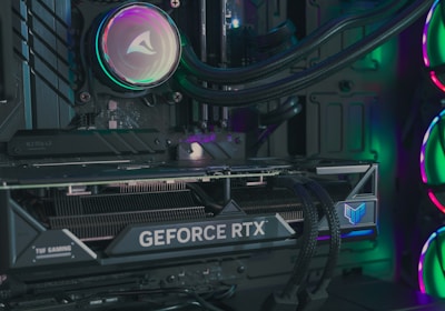 The image depicts the inside of a computer case featuring a high-performance graphics card labeled 'GEFORCE RTX' with RGB lighting. The cooling fans are illuminated with vibrant colors, creating a visually striking look. Various cables and components are neatly arranged, highlighting advanced hardware and a focus on aesthetics.