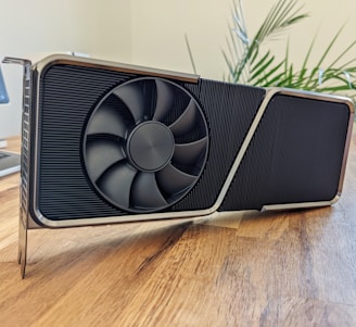 A large, modern graphics card with a bold design sitting on a wooden surface. The card features a prominent cooling fan and a sleek metal frame. In the background, a potted plant adds a touch of greenery and there is a partially visible computer monitor and keyboard.