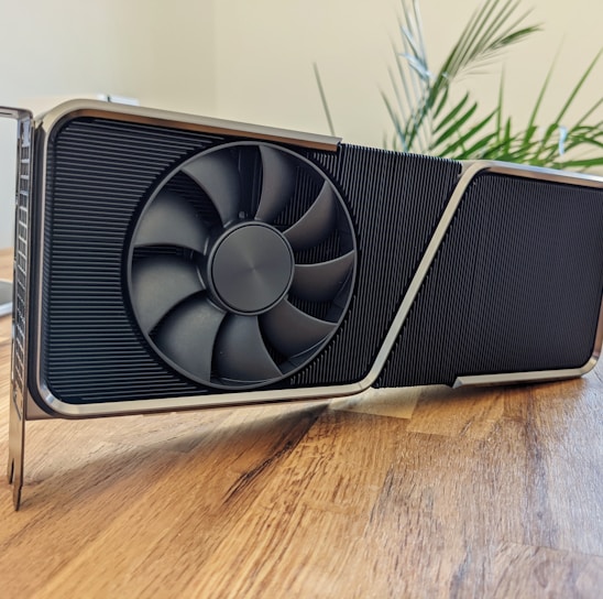 A large, modern graphics card with a bold design sitting on a wooden surface. The card features a prominent cooling fan and a sleek metal frame. In the background, a potted plant adds a touch of greenery and there is a partially visible computer monitor and keyboard.